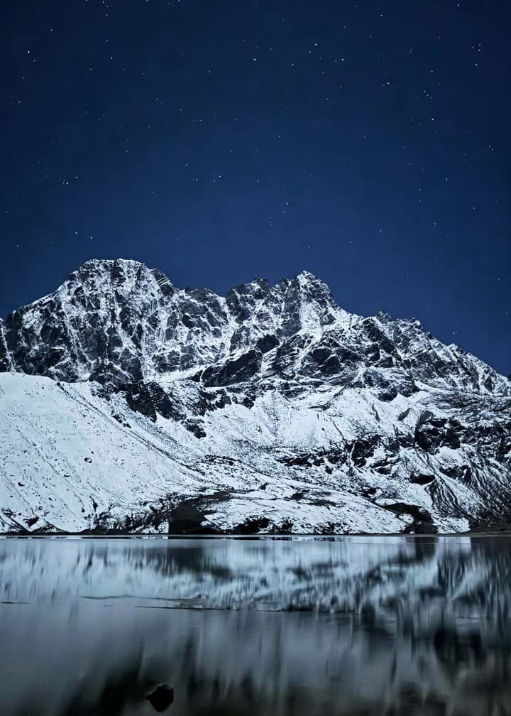 A snowy mountain reflecting on a dark lake under a starry sky.