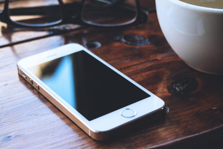 An older iPhone model sits on a wooden desk next to reading glasses and a cup of coffee.