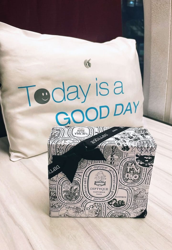 A pillow that reads "Today Is A Good Day" with a Diptyque toiletries set in a business class flight seat.