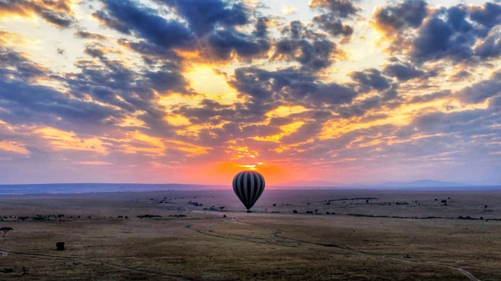 A hot air balloon at sunrise. The sky is orange and yellow, and the clouds are indigo and purple, over a yellow and brown flat landscape.