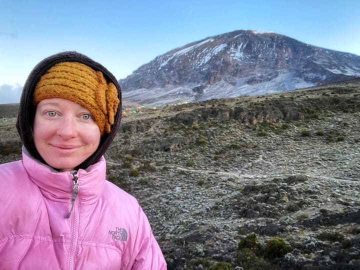 Clara Ritger poses in winter clothes with tiny tents seen far in the distance, in the shadow of the rounded Uhuru Peak, Mount Kilimanjaro's highest summit.