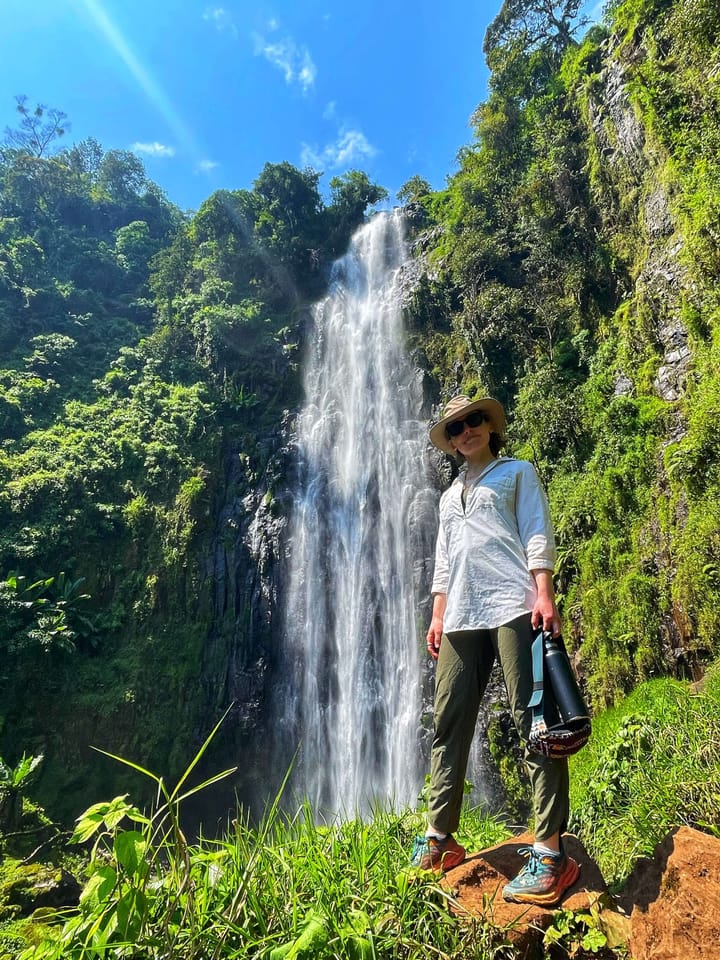 Clara Ritger stands next to a tall waterfall on a sunny, blue sky day.
