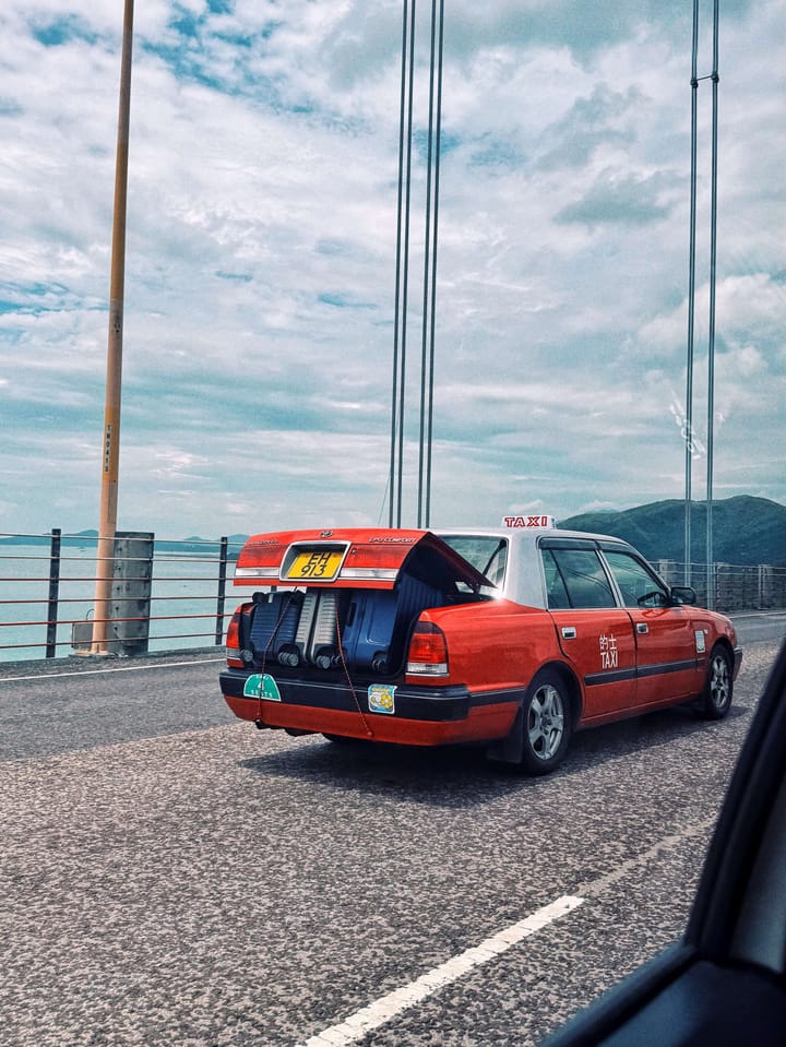 A red taxi in Hong Kong with an open trunk and three large suitcases that are hanging out the back. 