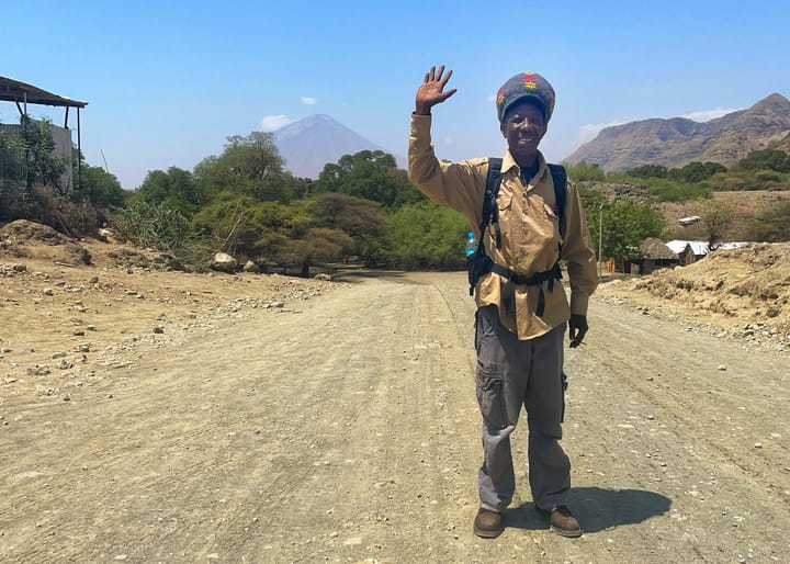 An African man in a khaki longsleeve and cargo pants waves on a dirt road.