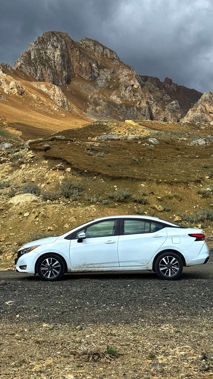 A small white car with mud tracks on it sits in the shadow of a rocky mountain. 