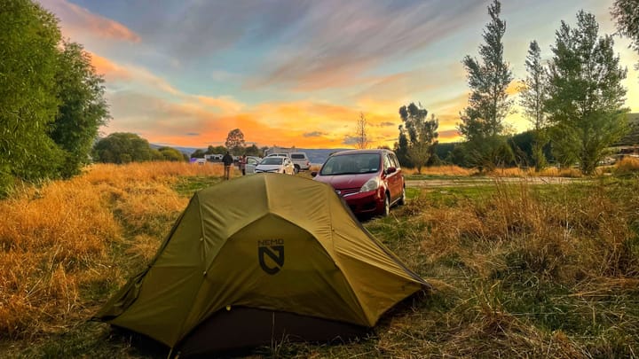 A green Nemo tent in front of a small red car, surrounded by tall grasses, and a sky painted with vibrant colors at sunset.