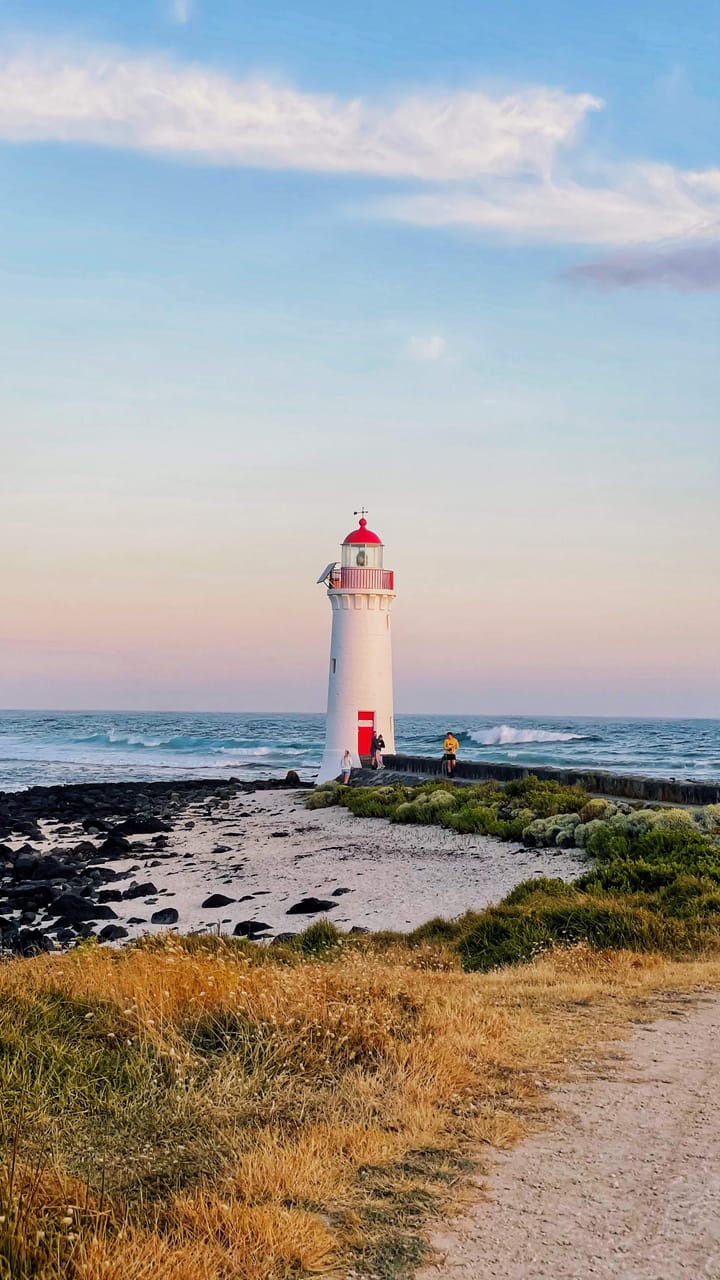 At white lighthouse with red embellishments sits at the edge of the ocean, with black lava rocks on a cream sandy beadh, and yellow grasses and green shrubbery lining a walking path. 